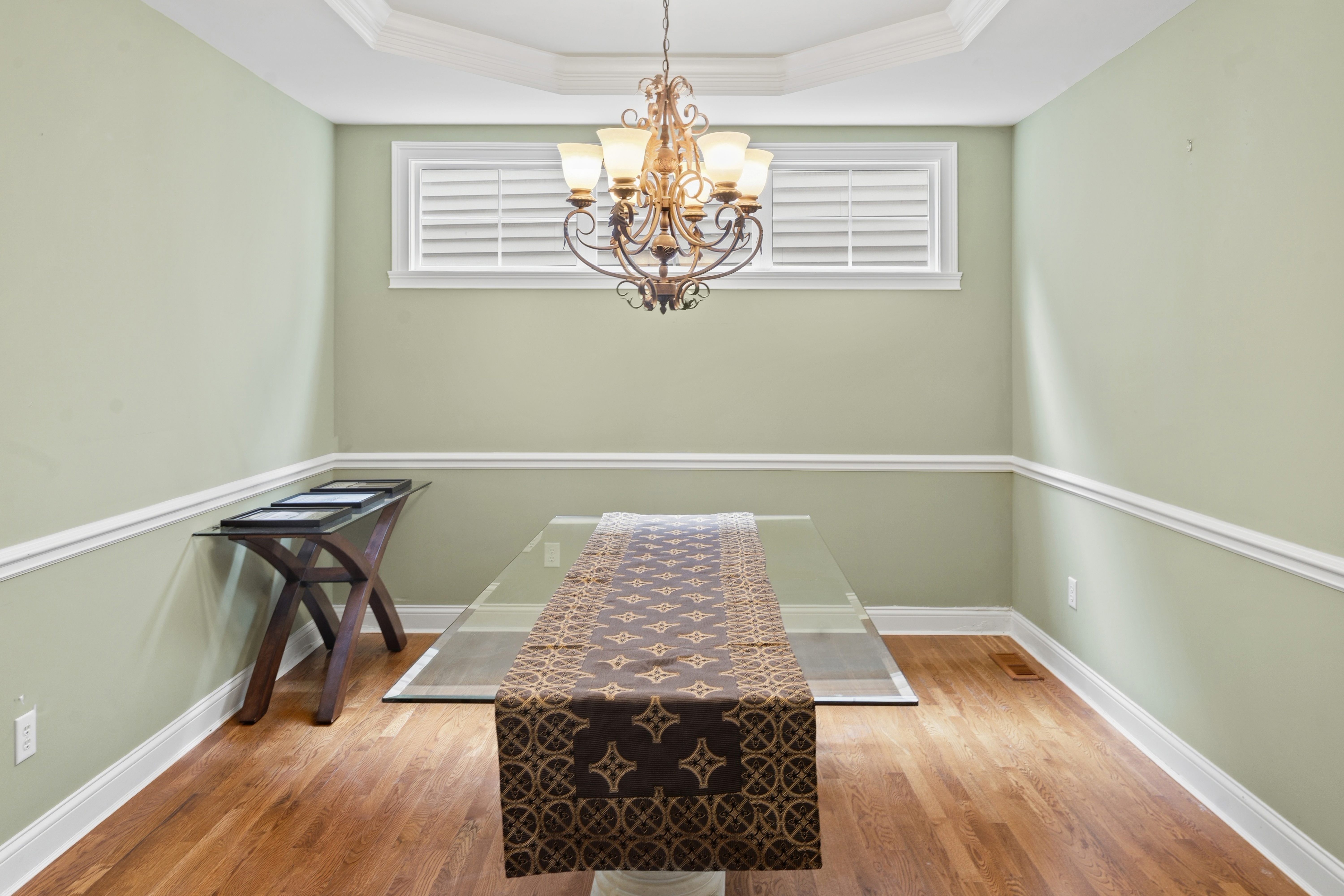 Formal dining room with tray ceiling, chandelier, glass dining table, and plantation shutters