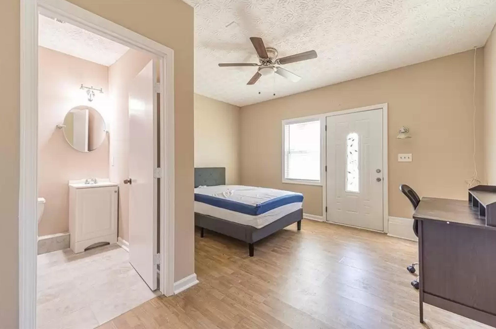 Bedroom with wood flooring, tan walls, ceiling fan, bed, desk, and attached bathroom view