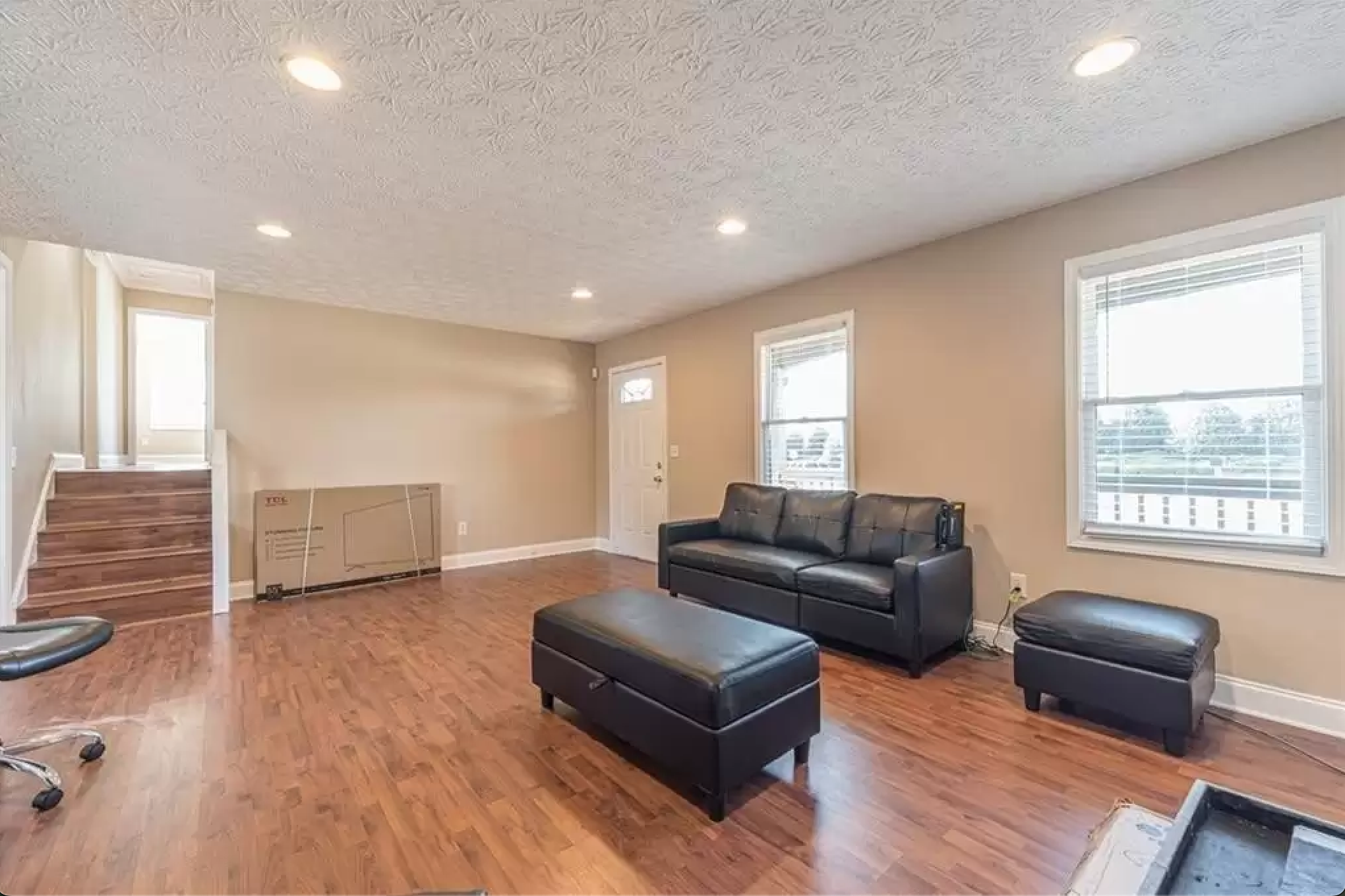 Living room with hardwood-style flooring, black sofa, ottoman, windows, and staircase